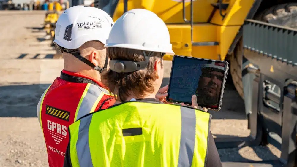 Two construction workers in high-visibility vests and hard hats reviewing a digital map on a tablet at a construction site, with GPRS branding visible.
