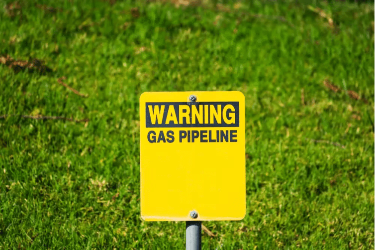 A yellow warning sign with the text 'WARNING GAS PIPELINE' in black letters, mounted on a metal post, set against a background of green grass.