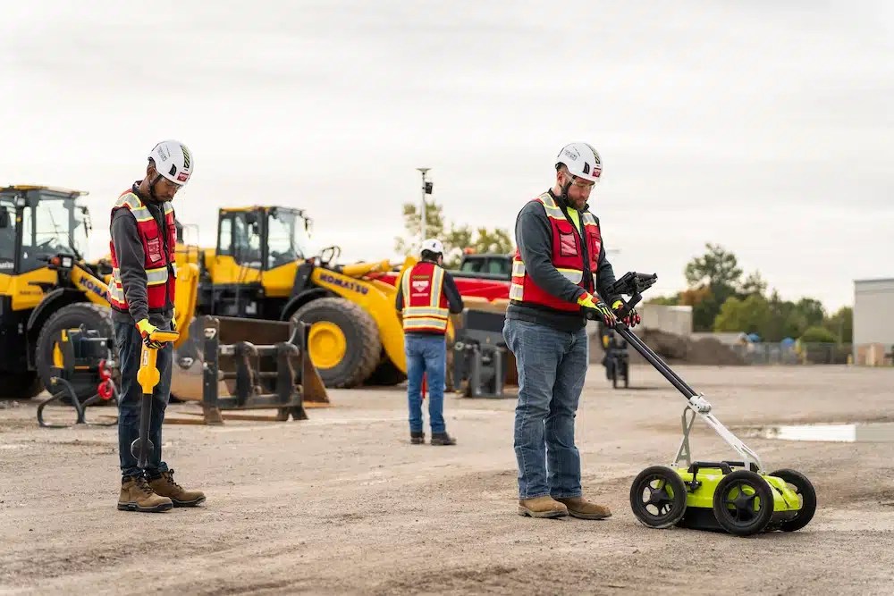 Three construction workers in safety gear operate utility locating equipment at a job site. One uses a handheld device while another pushes a ground-penetrating radar (GPR) unit. Yellow construction vehicles and equipment are visible in the background under an overcast sky.