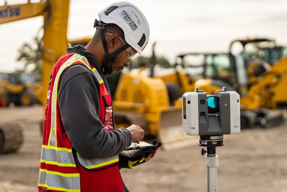 GPRS Project Manager in safety gear operating a 3D laser scanner at an active construction site, with heavy machinery in the background.