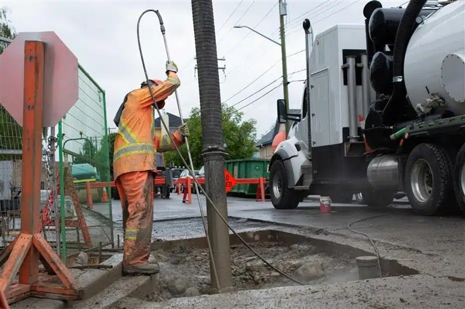 A worker using a hydro vac system to excavate under pavement in a city.