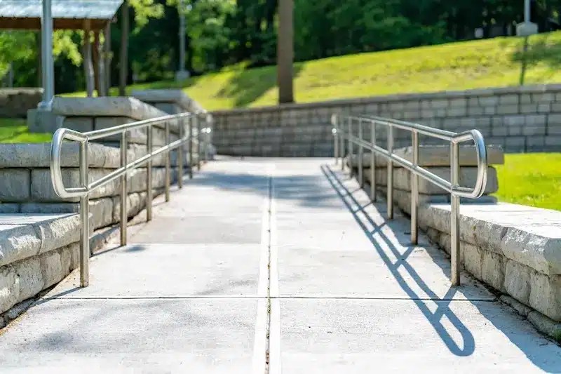 Concrete pathway with metal handrails on both sides, leading to a grassy area with trees and a stone retaining wall in the background.