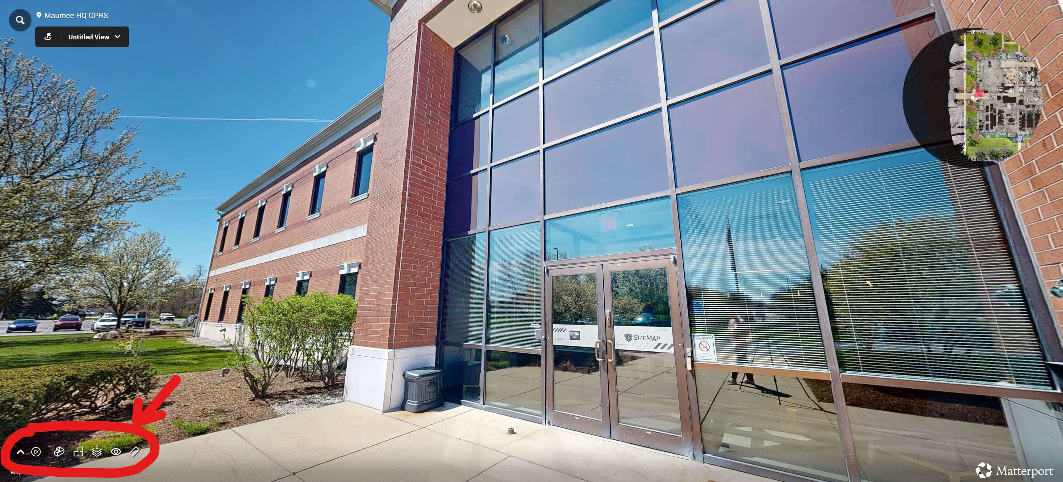 Exterior view of a modern brick building with large reflective glass windows, captured in a 3D virtual walkthrough interface. A small aerial map is shown in the top-right corner, and interactive navigation tools are highlighted in the bottom-left corner. 