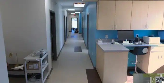 Hospital corridor with blue accent wall, nurse station with computer and cabinets, exam room doors, a medical cart on the left, and daylight at the far end.