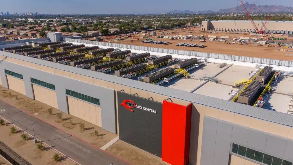 Aerial view of a large data center facility with rooftop cooling equipment, surrounding infrastructure, and nearby construction activity.