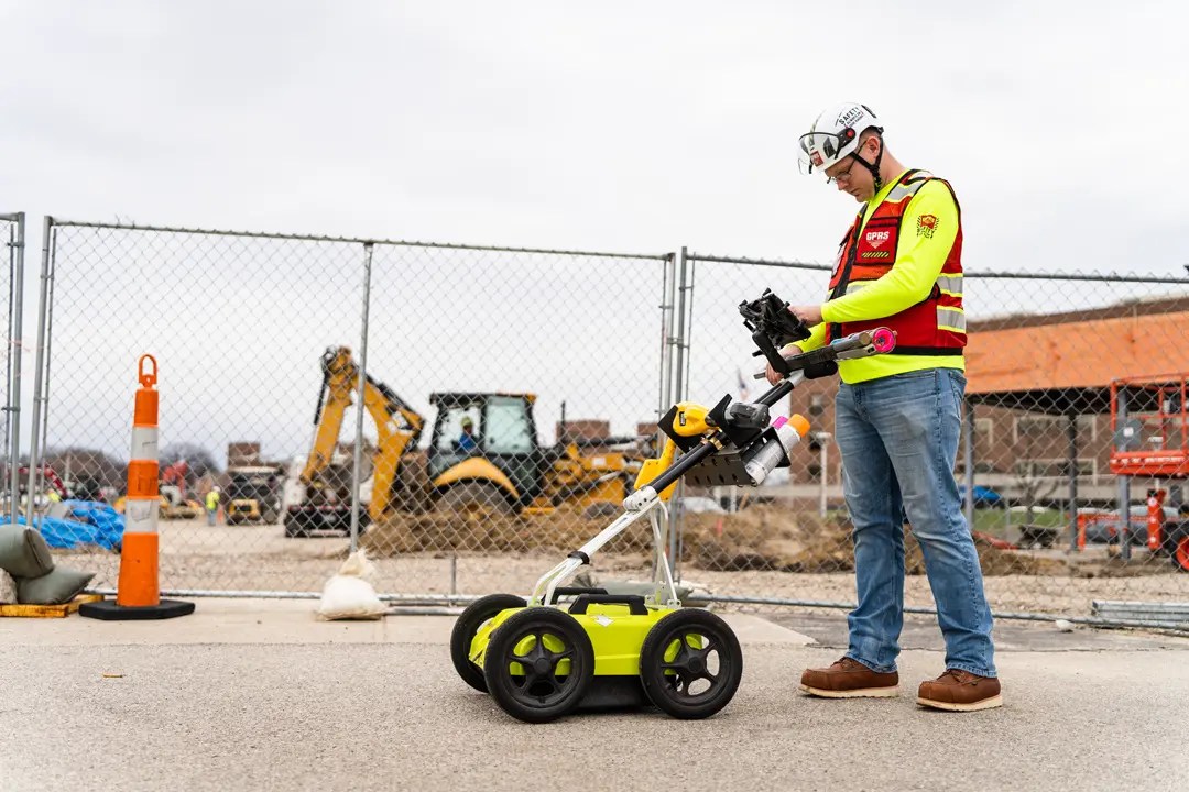 GPRS Project Manager using ground penetrating radar equipment to locate underground utilities at an active construction site behind a security fence.