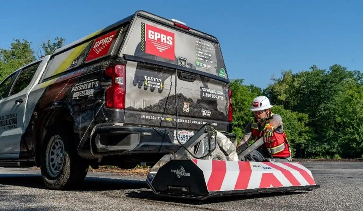 A GPRS-branded truck is parked on a paved surface with a high‑speed ground‑penetrating radar (GPR) array positioned behind it. A GPRS field team member wearing a hard hat and high‑visibility vest is kneeling beside the GPR array, preparing it for use. Trees and greenery line the background.