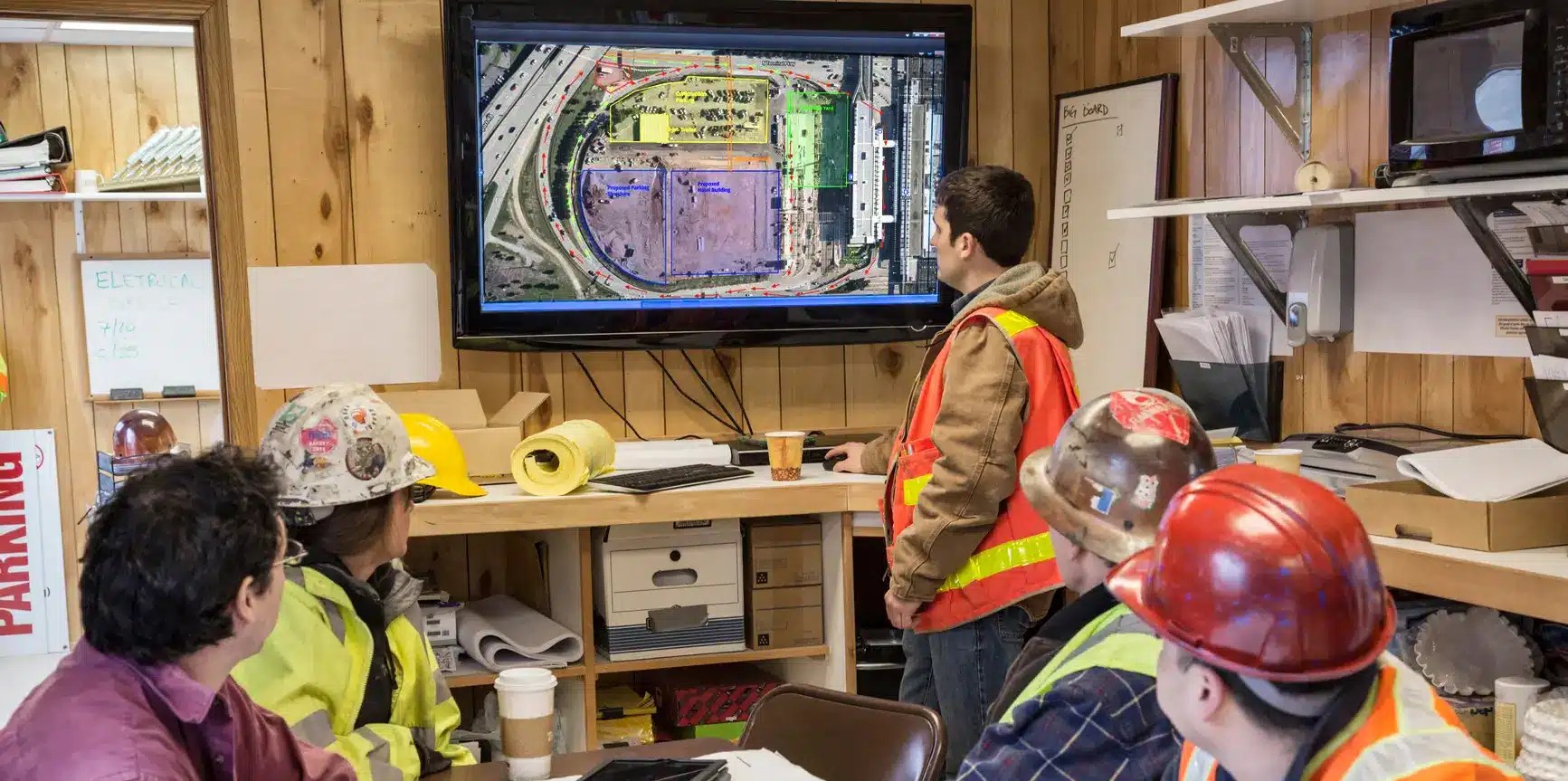 Construction workers seated around a table in a jobsite trailer while a digital construction site map is displayed on a large screen.