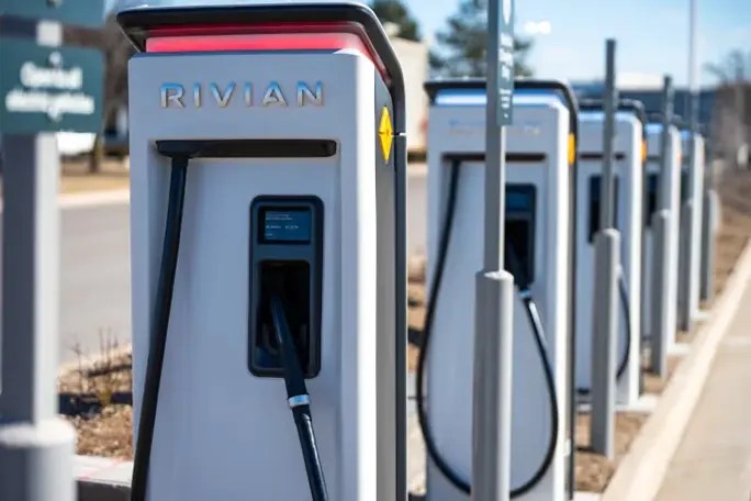 Close-up of Rivian electric vehicle charging stations lined up along a parking lot curb, with charging cables connected and additional chargers visible in the background.