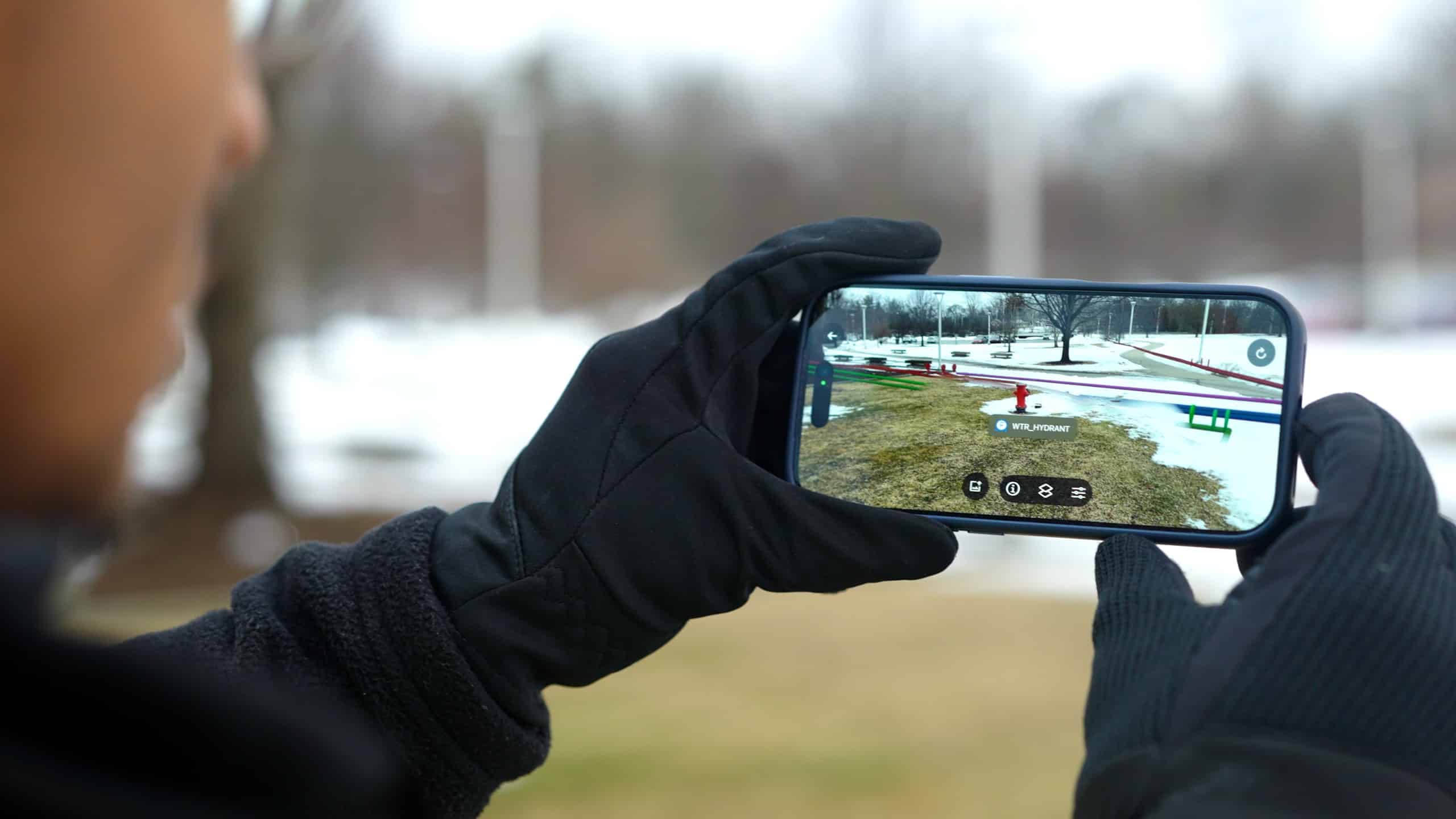 Close-up of a person wearing black gloves holding a smartphone outdoors, displaying a SiteMap augmented reality map overlay with colored markers and icons on a grassy area with patches of snow, with a blurred winter landscape in the background.