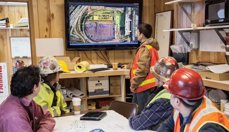 Five construction workers wearing hard hats and safety vests sit around a table in a wood-paneled site office while one worker stands in front of a wall-mounted screen displaying a SiteMap software aerial map with marked utilities and boundaries; blueprints, coffee cups, and notebooks are spread across the table as the group looks toward the screen.